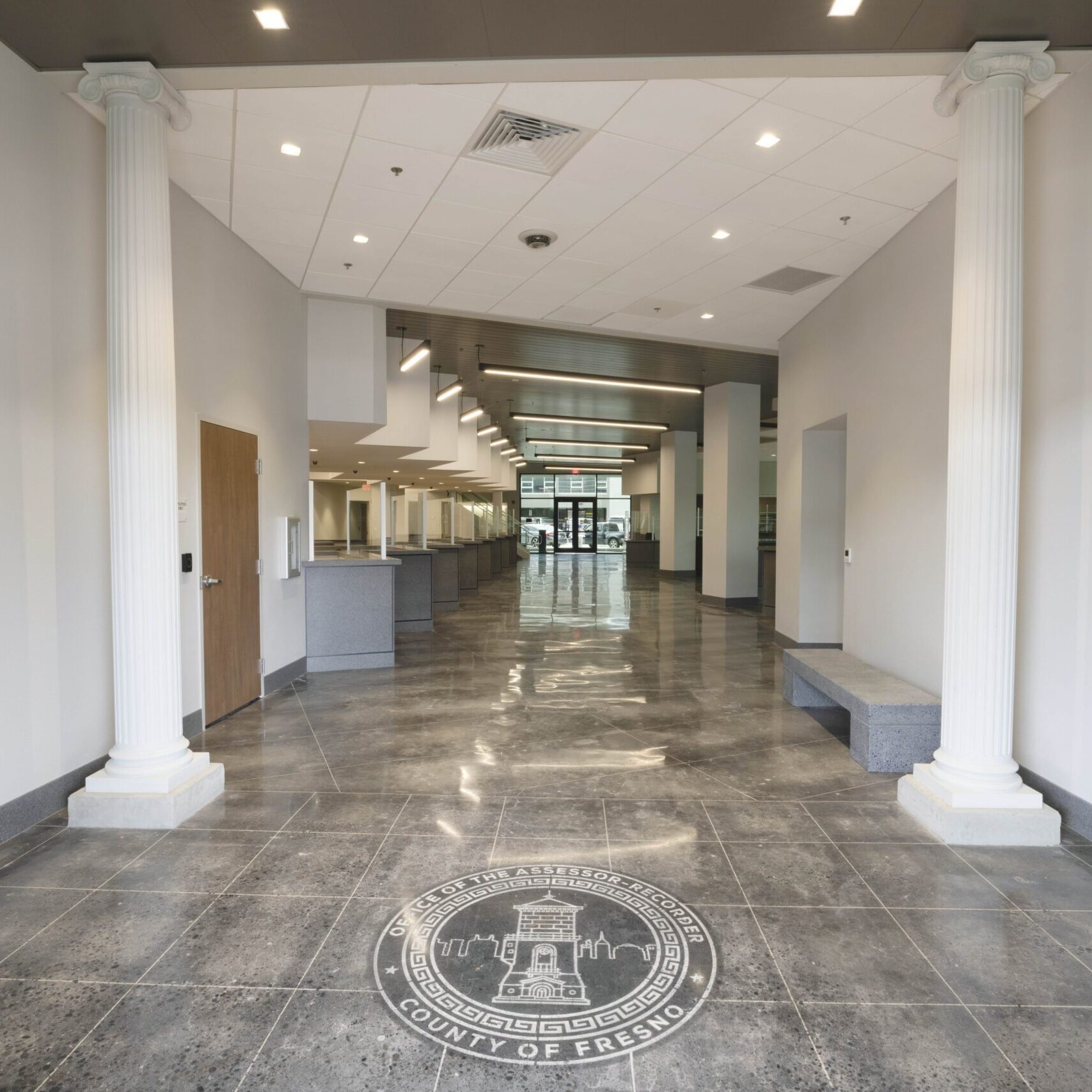 A low-angle, eye-level shot from inside a large building, looking out through the front doors. The floor is a dark, reflective tile with a large, circular seal of Fresno County in the center. Two large, white columns with a fluted pattern flank the entrance. The hallway leads to a long series of service counters in the background. The room has a high ceiling with bright recessed lighting.