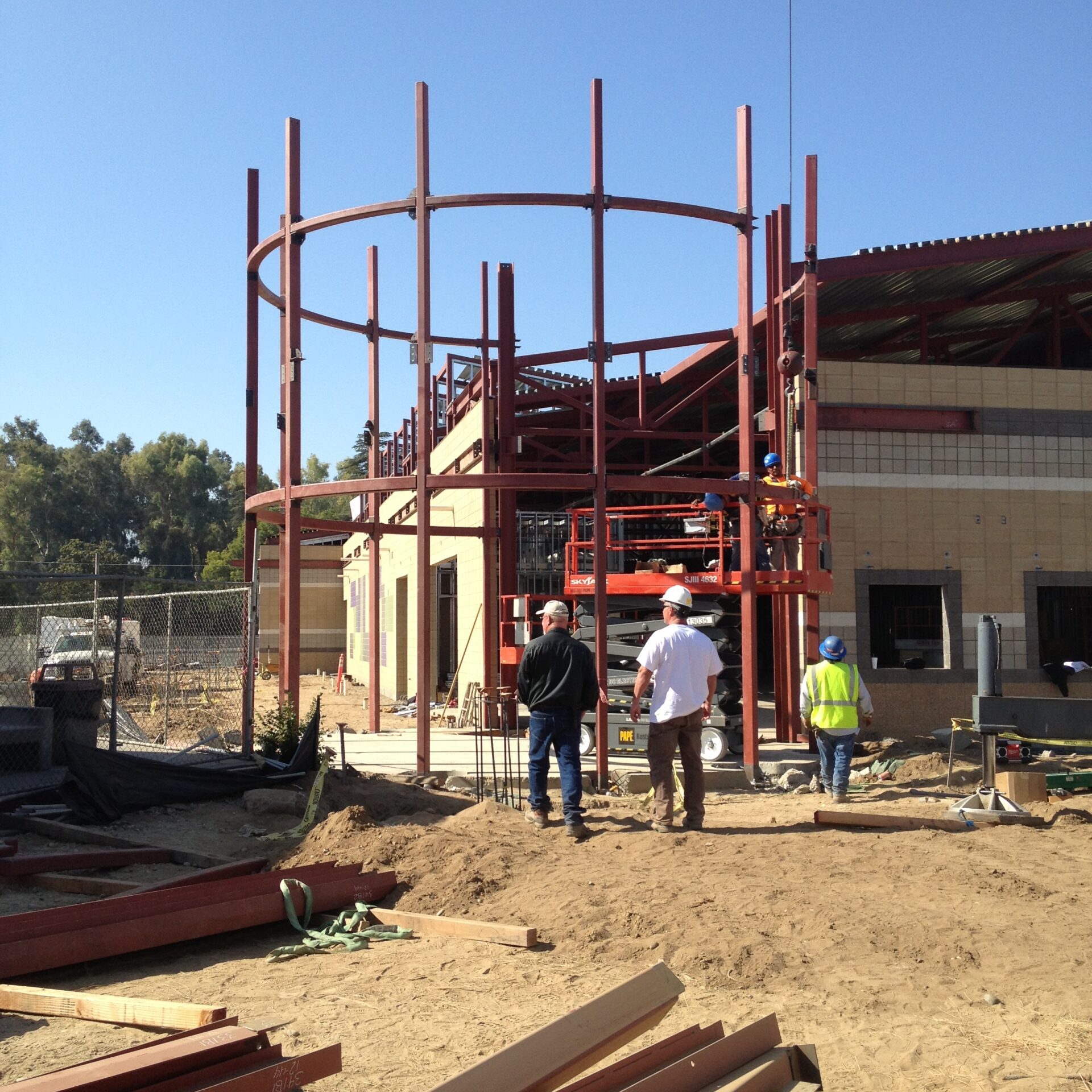 A group of construction workers stand on a dirt lot, observing the steel framing of a circular structure that will form the entrance of a building. The main building, with its exposed roof trusses, is visible behind the circular frame. A scissor lift is positioned at the back of the circular structure, and loose steel beams are scattered on the ground in the foreground.