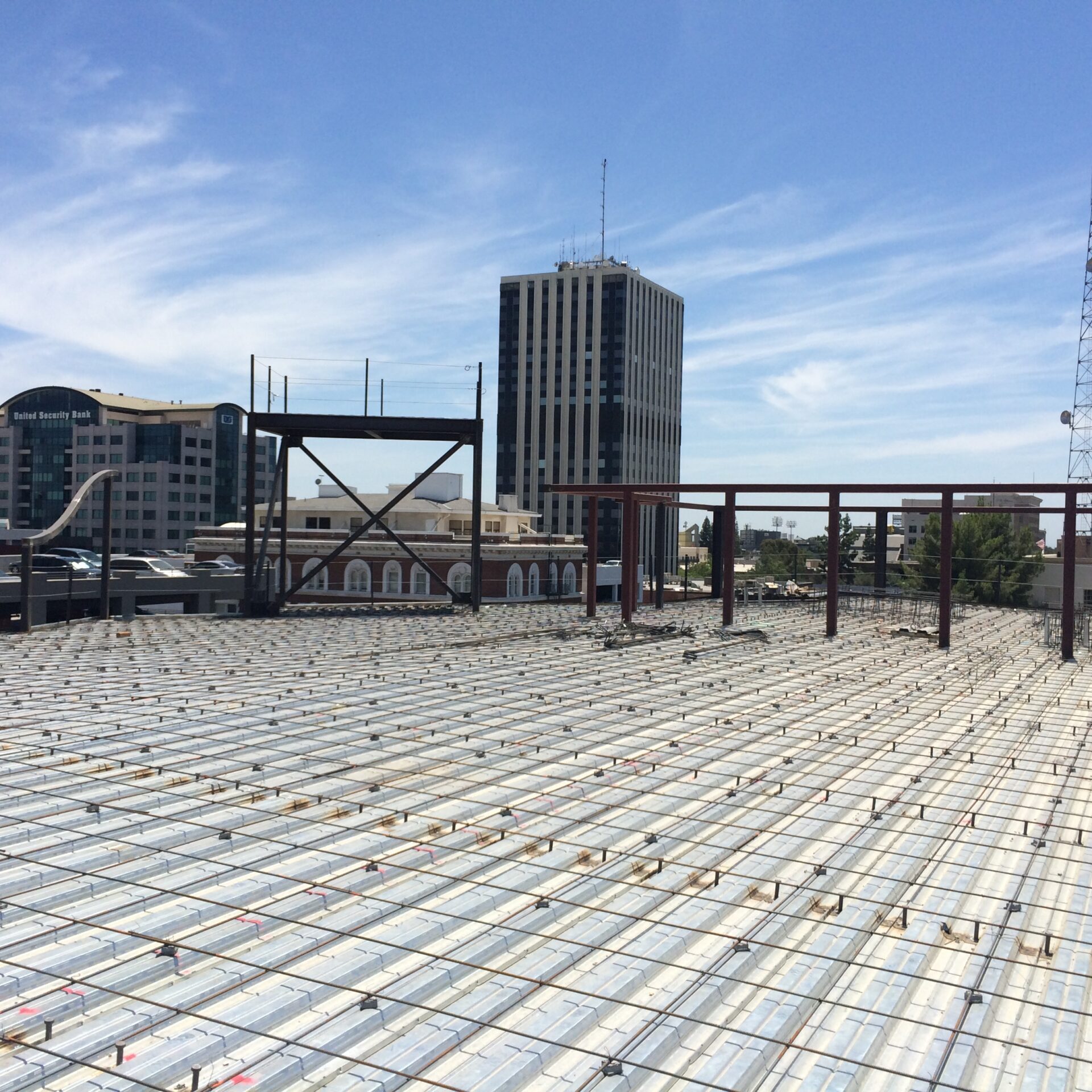 A low-angle, eye-level shot from a rooftop under construction, looking out at the Fresno, California skyline. In the foreground, the roof is covered with a grid of metal decking and rebar. A large steel framework and scaffolding are visible in the middle ground, with a parking garage to the left. In the background, there are two tall buildings and a large red-brick building with arched windows. A tall, gray cell tower stands on the right. The sky is blue with scattered cirrus clouds.