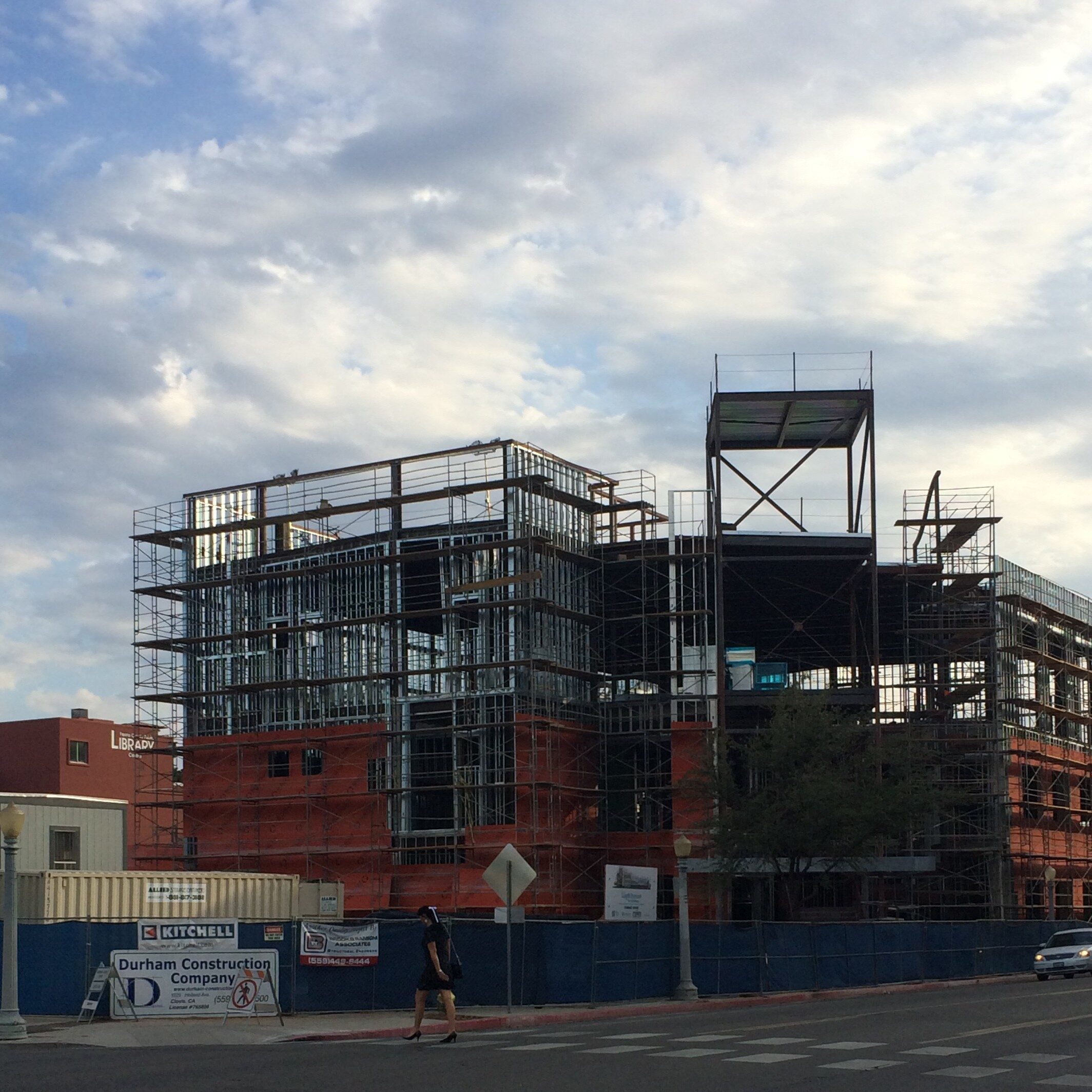 A multi-story building is under construction, with its exposed steel frame and scaffolding visible against a cloudy sky. The structure is partially covered in an orange, mesh-like material. A sign for Durham Construction Company is posted on a blue fence in the foreground. A person is walking across the street on a crosswalk in front of the construction site.