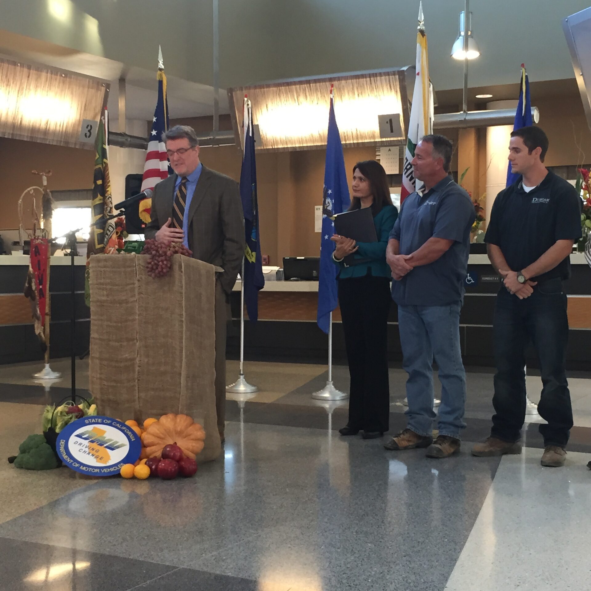 A group of four people, three men and one woman, are standing inside a modern building with high ceilings and polished floors. The main speaker, a man in a suit, is at a podium decorated with fresh gourds, fruits, and a sign for the State of California Department of Motor Vehicles. Behind them, several flags, including the American flag and the California state flag, are visible, along with multiple service counters.
