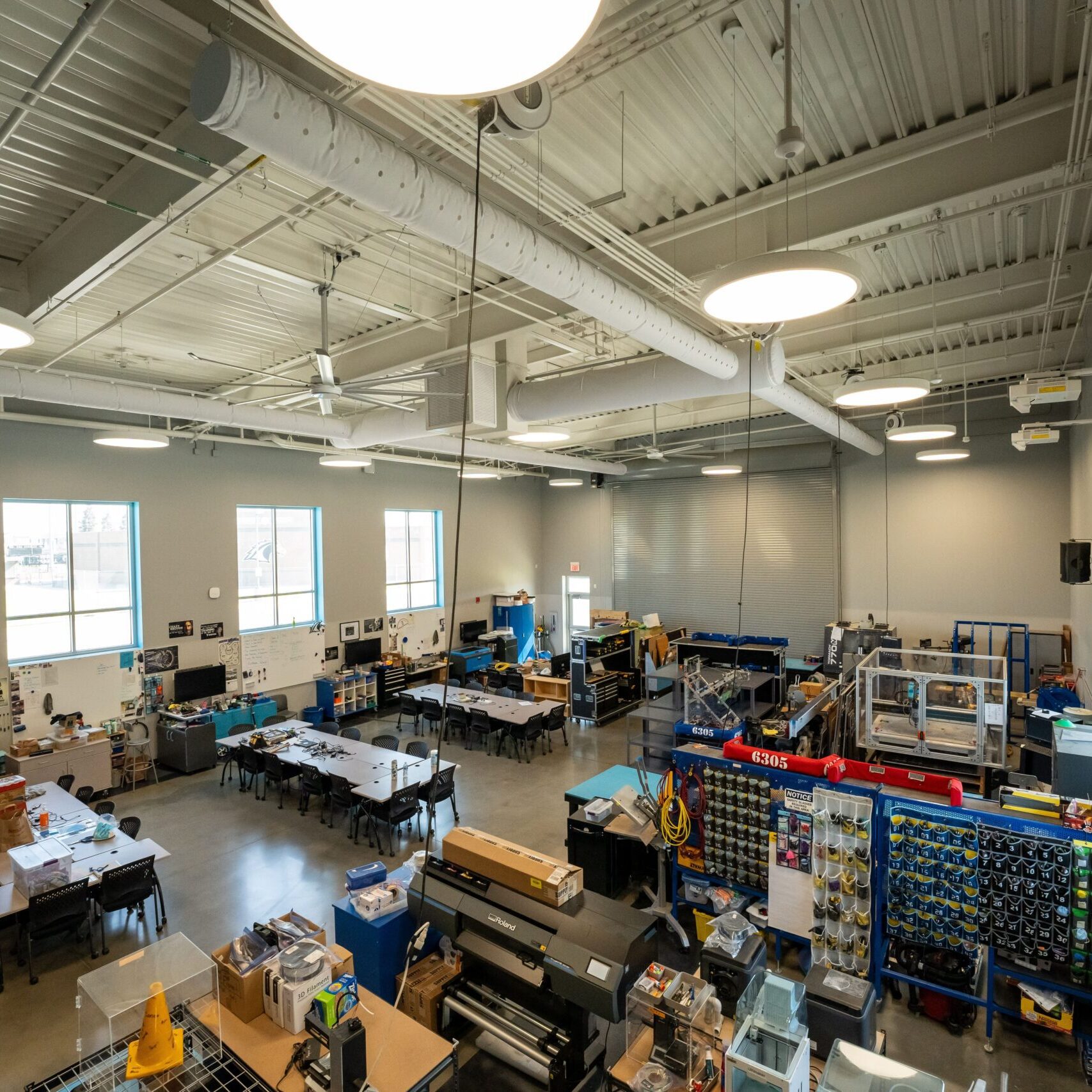 A wide-angle, elevated interior shot of a workshop. The room is spacious with a high ceiling and concrete floors. Various worktables and equipment, including a large industrial-looking machine and a storage rack filled with parts, are visible. The walls have large windows and a mural.