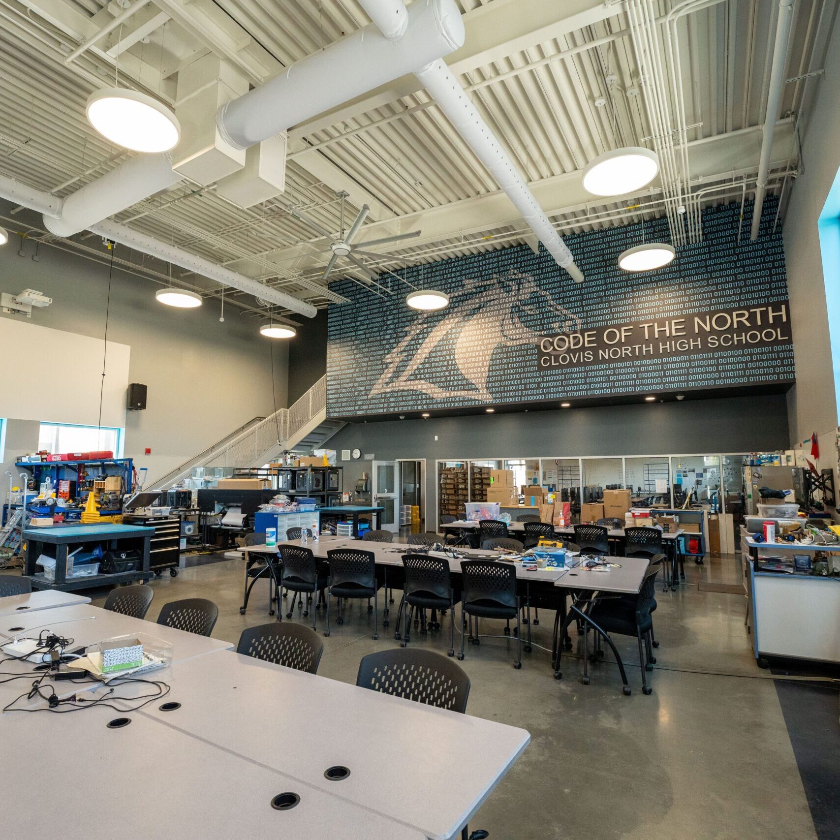 An interior view of a high-ceilinged workshop space. A large mural with a binary code pattern and the words "CODE OF THE NORTH CLOVIS NORTH HIGH SCHOOL" is on the back wall. Worktables, chairs, and various robotics equipment are arranged in the room.