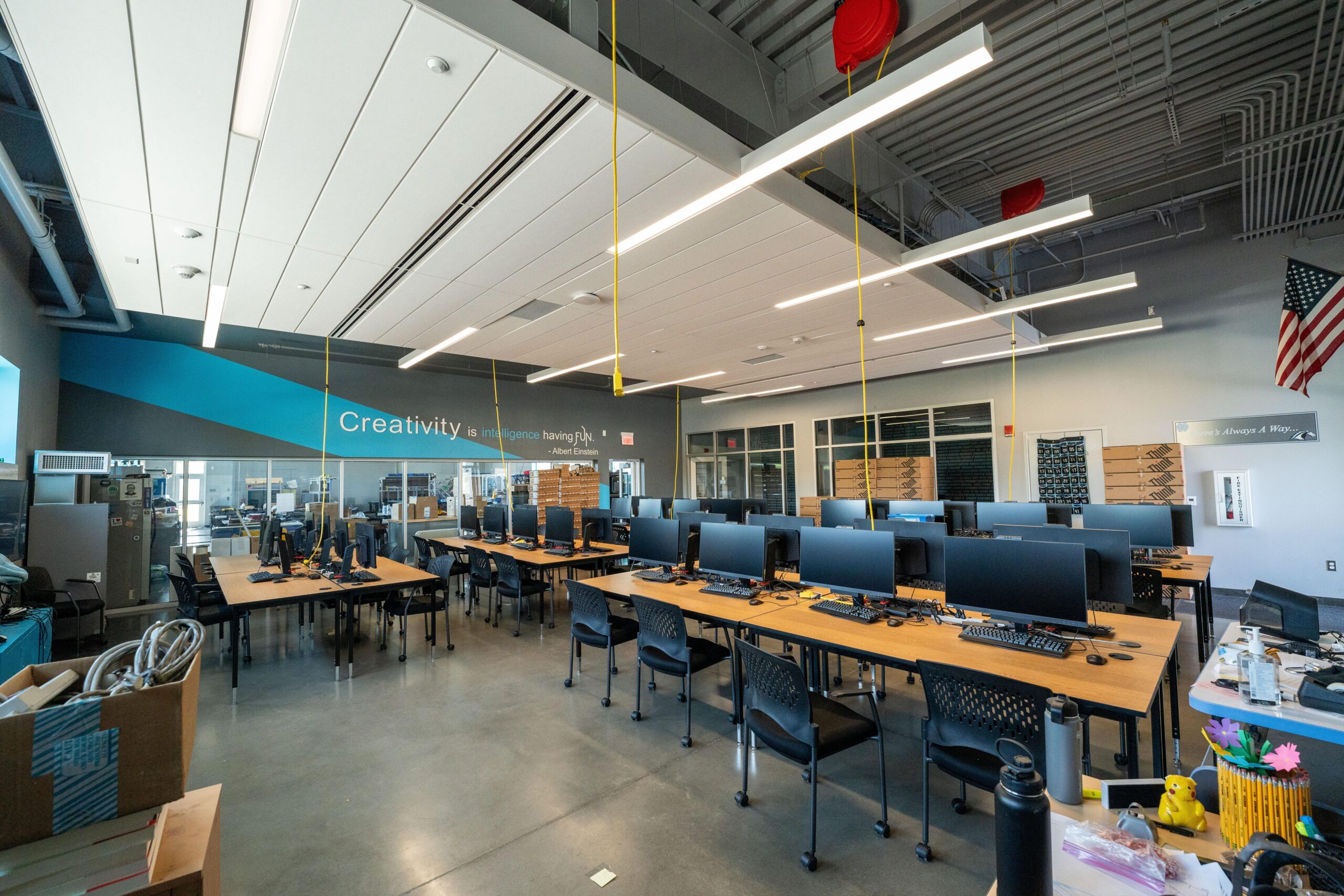 Interior (3 of 14)-min A wide-angle interior shot of a modern, open-plan classroom or computer lab. Rows of long wooden desks with black chairs are arranged in the room, each with a desktop computer monitor and keyboard. A blue and gray accent wall in the back reads "Creativity is intelligence having fun." The ceiling has exposed ductwork, pipes, and modern linear light fixtures hanging from the ceiling. A US flag is hanging on the far right wall. There are large windows looking into an adjacent room. The floor is a polished concrete surface. In the foreground, a desk is cluttered with a water bottle and various items.