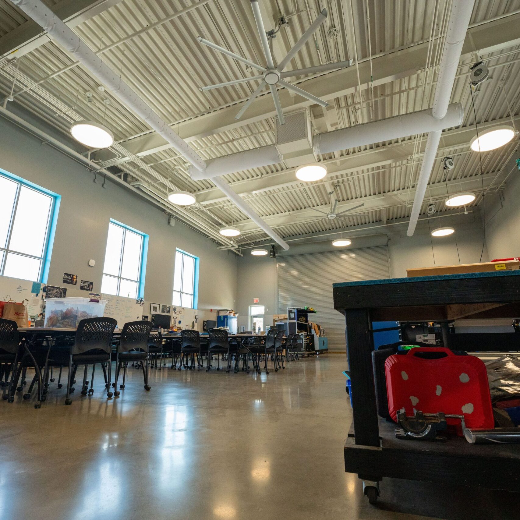 An interior view of a modern classroom or workshop. The room has high ceilings, polished concrete floors, and large windows. Multiple desks with chairs are arranged in the center, and various tools and materials are on the workbenches.