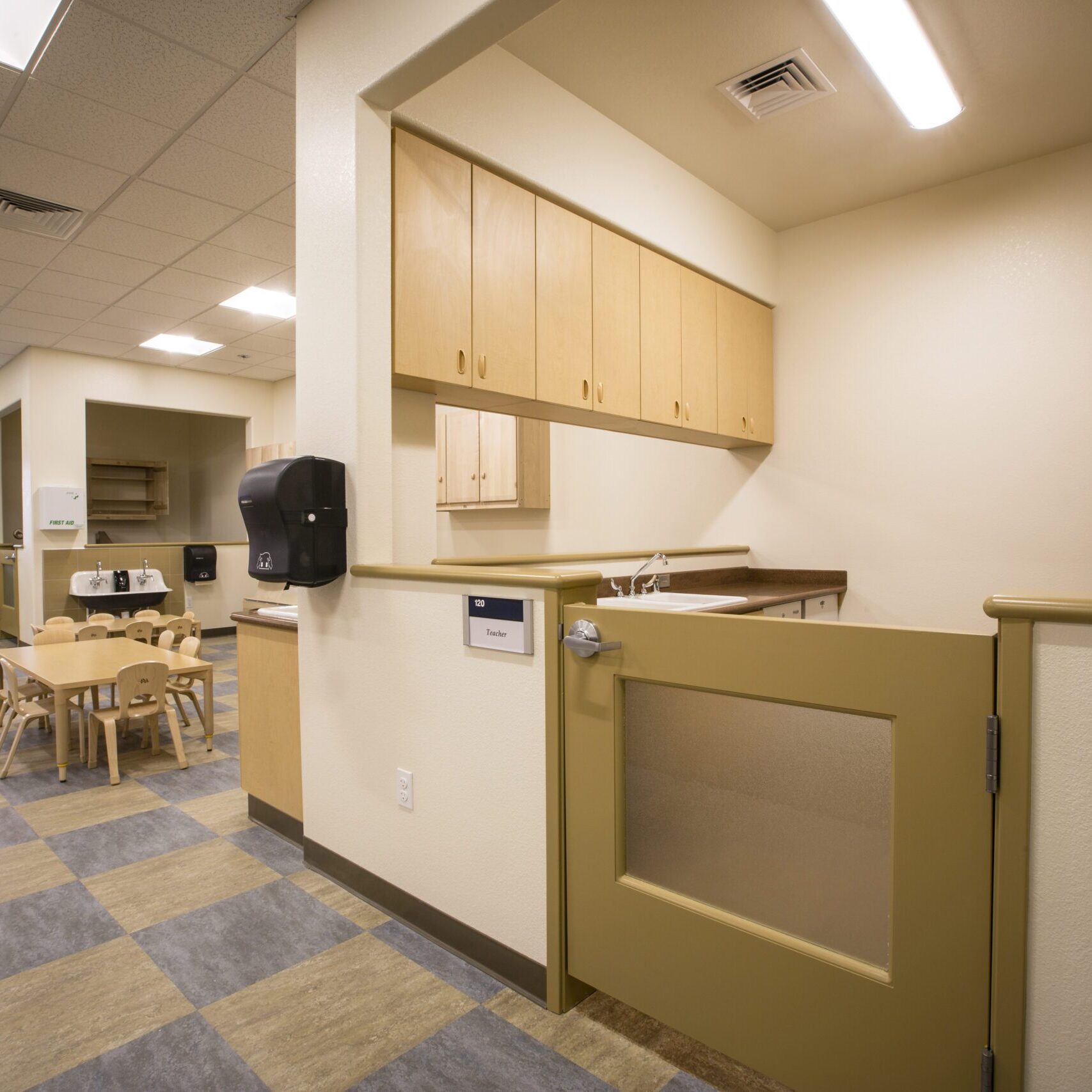 A wide-angle, low-shot of a bright, clean classroom or play area. In the foreground, there is a half-height door with a frosted-glass panel that leads to a small kitchenette area. The kitchenette has a sink, countertop, and light-wood cabinets. The area is marked with a sign that reads "Kitchen" and is lit by a long fluorescent light. In the background, a small wooden table with several chairs is visible on a blue and tan checkered floor. The walls are a light beige, and the ceiling has square tiles with recessed lighting.