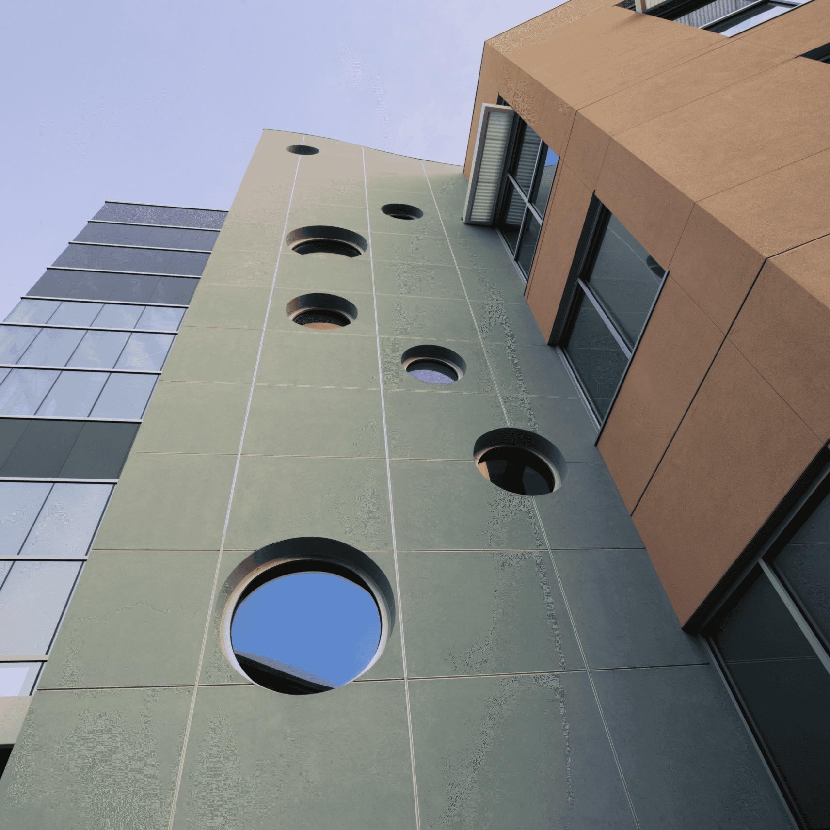 A low-angle shot of a modern building with two distinct facades. On the left, a glass facade reflects the sky and a nearby structure. The central section is a muted green-gray color with a series of decreasingly smaller circular windows, with the largest one at the bottom and a rectangular vent above. To the right, a tan-colored, flat-paneled section of the building is visible with large rectangular windows. The different textures and shapes of the building are shown against a clear blue sky.