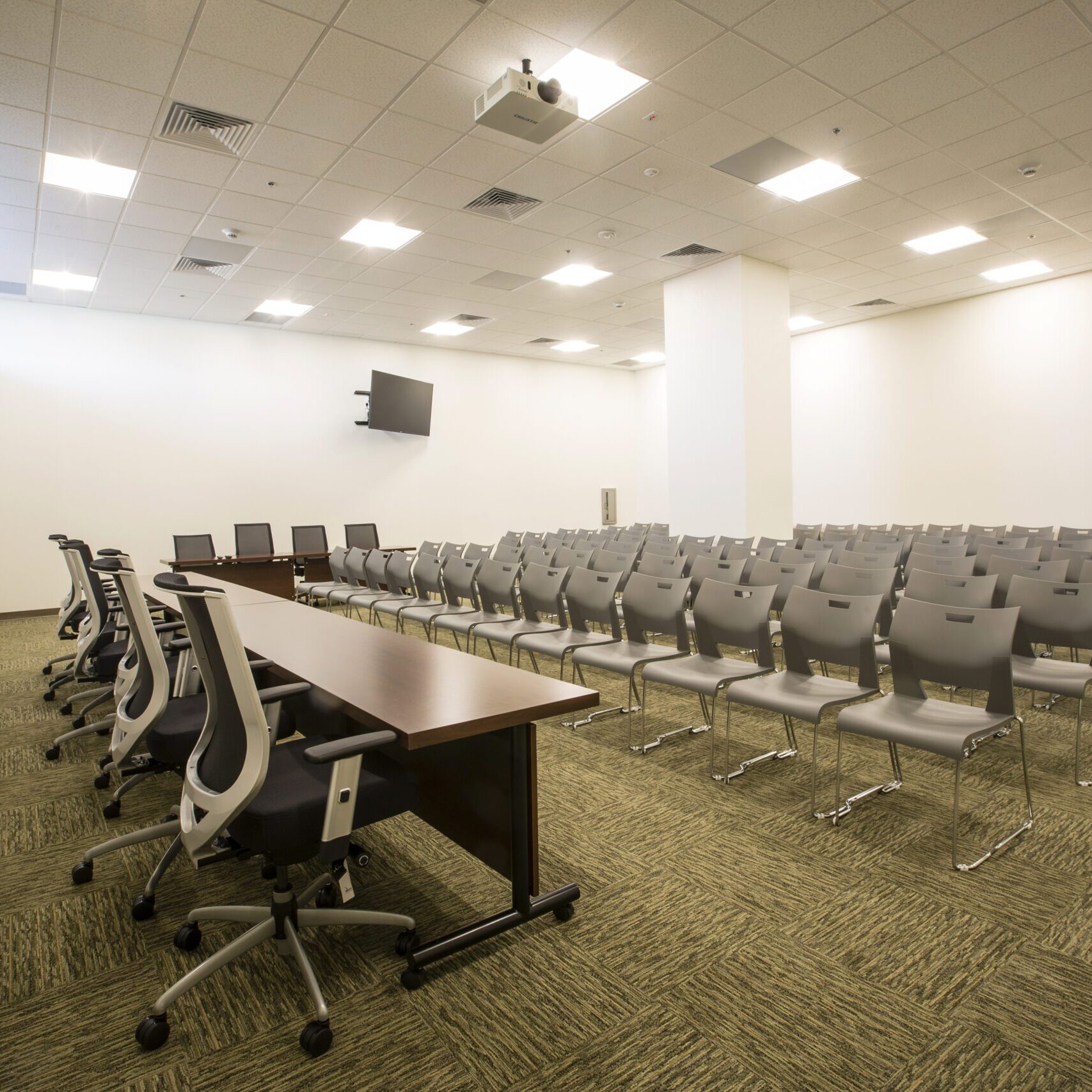 A large, empty conference room or auditorium with a low, wide-angle shot. In the foreground, there is a long, dark wood conference table with several black rolling office chairs arranged on either side. A long line of identical gray chairs is neatly arranged in rows behind the table. A projector is mounted on the ceiling, and a television screen is mounted on the back wall. The room has a beige and green patterned carpet, white walls, and bright overhead lights.
