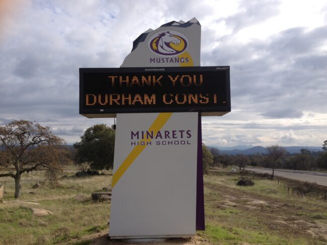A low-angle, exterior shot of the electronic marquee sign for Minarets High School. The sign is in a grassy field with trees and a road visible behind it. The top of the sign features the school's "Mustangs" mascot logo. The electronic display screen reads "THANK YOU DURHAM CONST.". Below the screen, the sign has the text "MINARETS HIGH SCHOOL" in a modern font with a yellow and purple design. The sky is filled with white and gray clouds. In the distance, hills and mountains are visible.