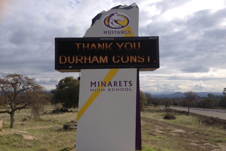 A low-angle, exterior shot of the electronic marquee sign for Minarets High School. The sign is in a grassy field with trees and a road visible behind it. The top of the sign features the school's "Mustangs" mascot logo. The electronic display screen reads "THANK YOU DURHAM CONST.". Below the screen, the sign has the text "MINARETS HIGH SCHOOL" in a modern font with a yellow and purple design. The sky is filled with white and gray clouds. In the distance, hills and mountains are visible.