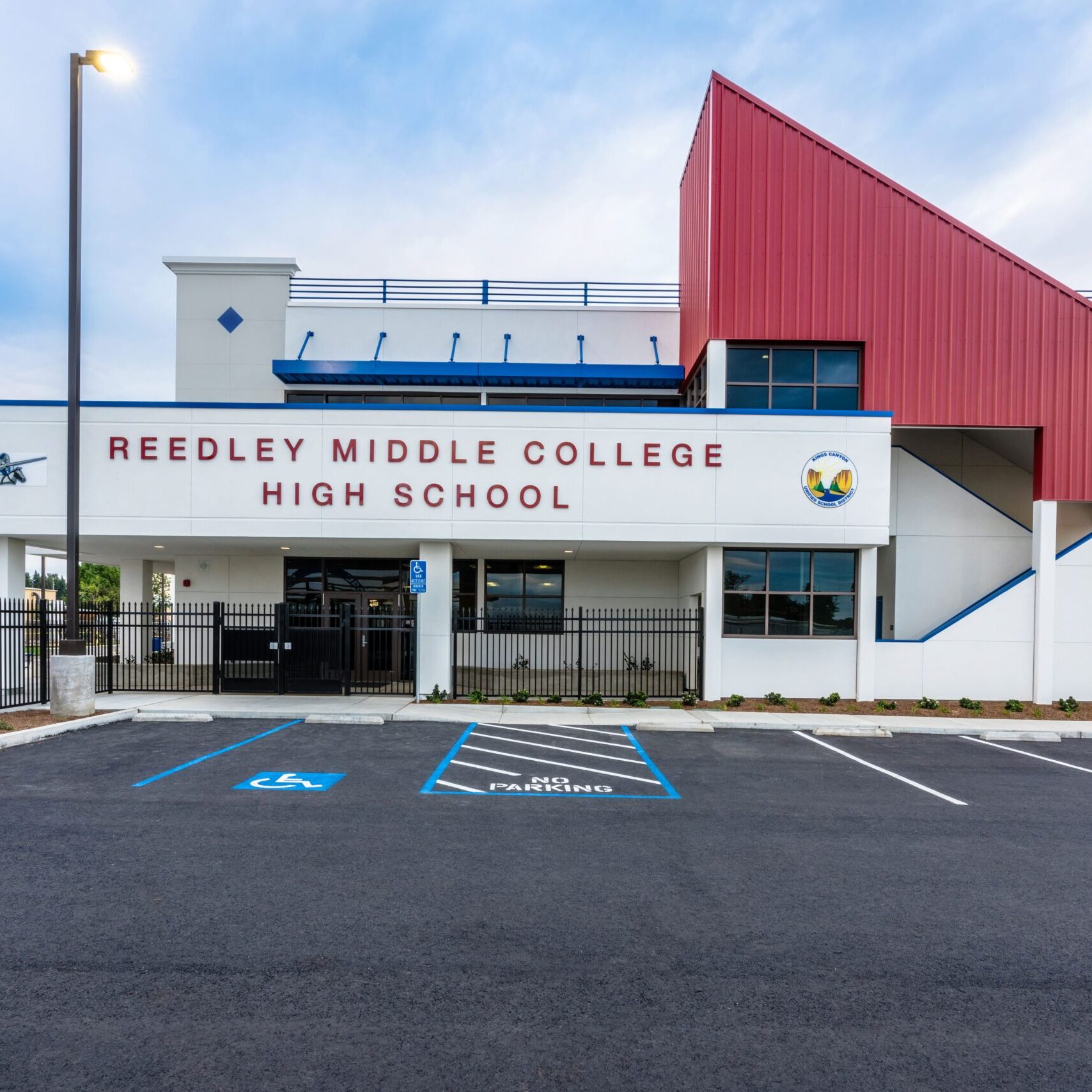 A front view of the modern Reedley Middle College High School building. The sign "REEDLEY MIDDLE COLLEGE HIGH SCHOOL" is clearly visible in a maroon font on a white wall. The building has a red angled roof on the right and a blue and white roofline on the left.