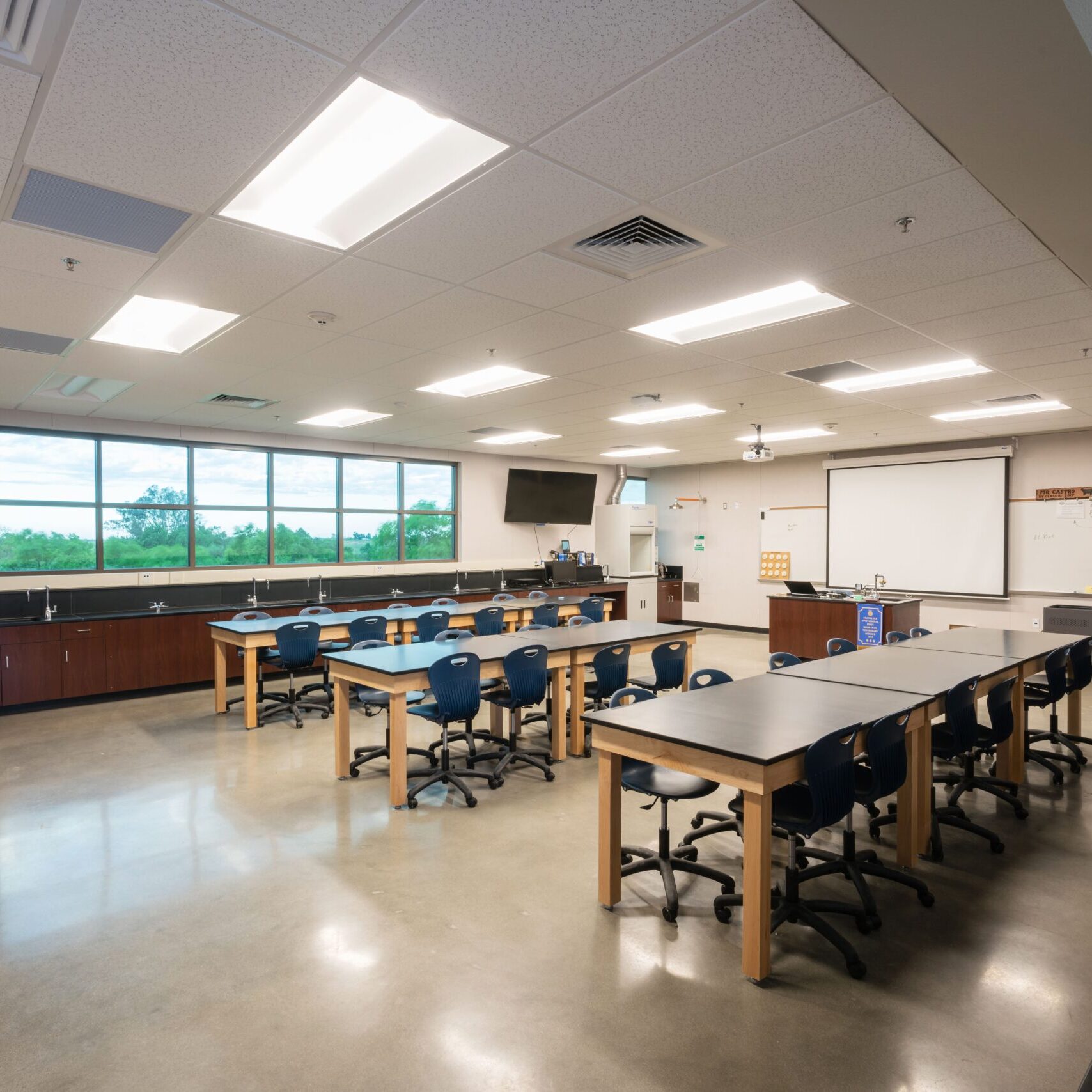 An interior shot of a new science classroom. The room has multiple black lab tables with stools, a long countertop with sinks along the far wall, and a large window. A projector screen is lowered near the front of the room.