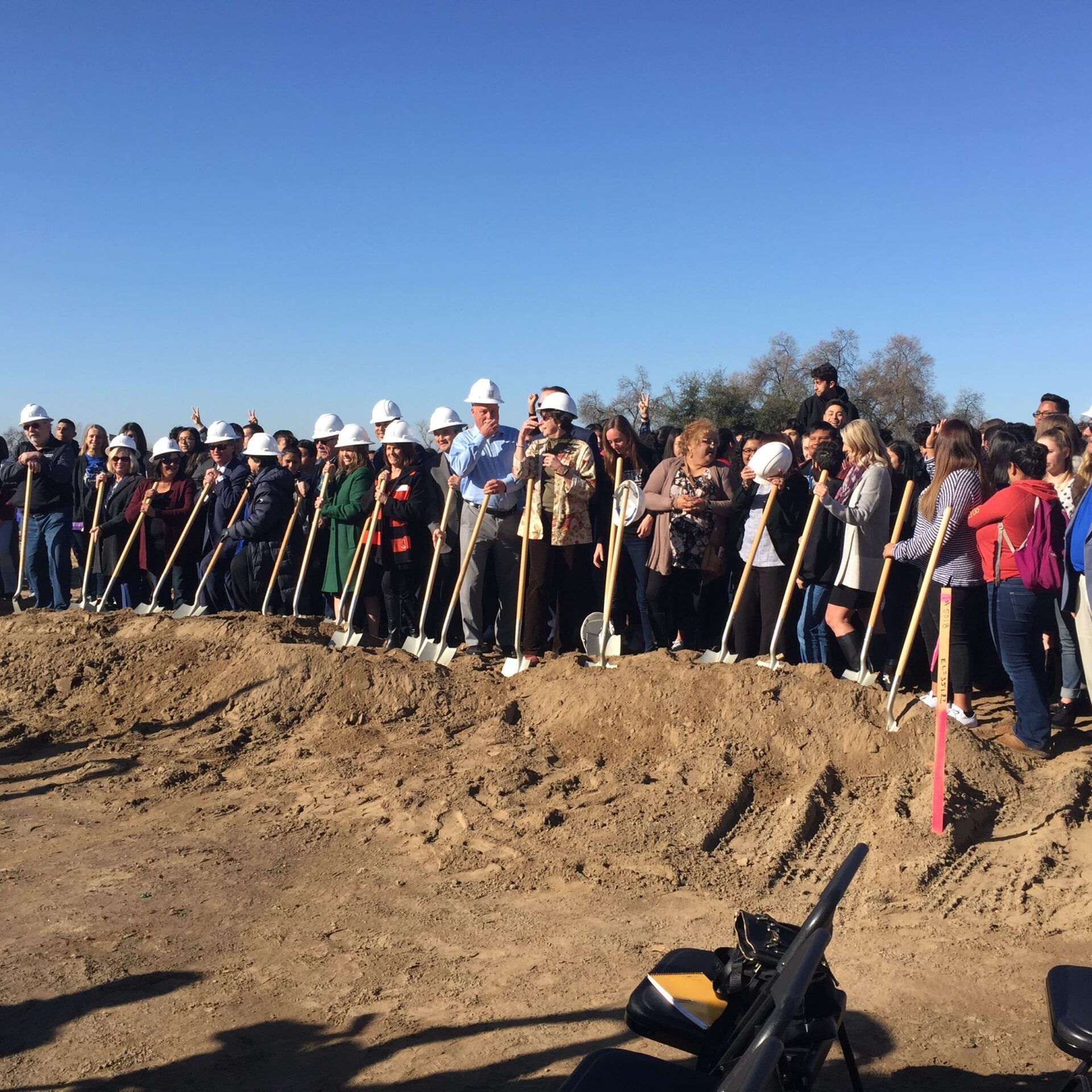 A group of people, many wearing white hard hats, are lined up behind a dirt mound, holding shovels. The event appears to be a groundbreaking ceremony for the Reedley Middle College High School.