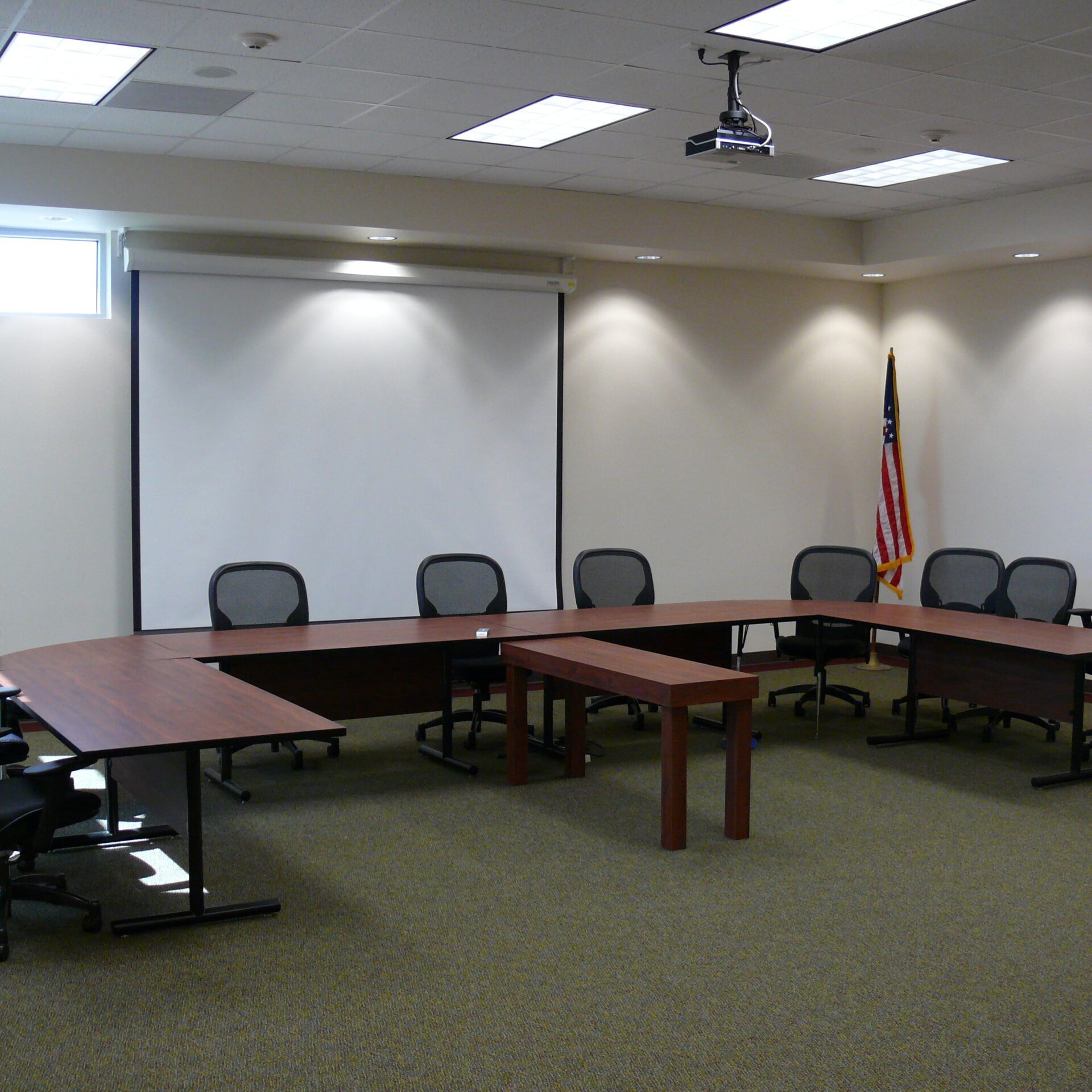 The interior of a modern conference room. A large, U-shaped wooden table is surrounded by multiple rolling office chairs. A projector screen is at the head of the table, and an American flag stands in the corner. The room is brightly lit with recessed ceiling lights and has a neutral-colored carpet.