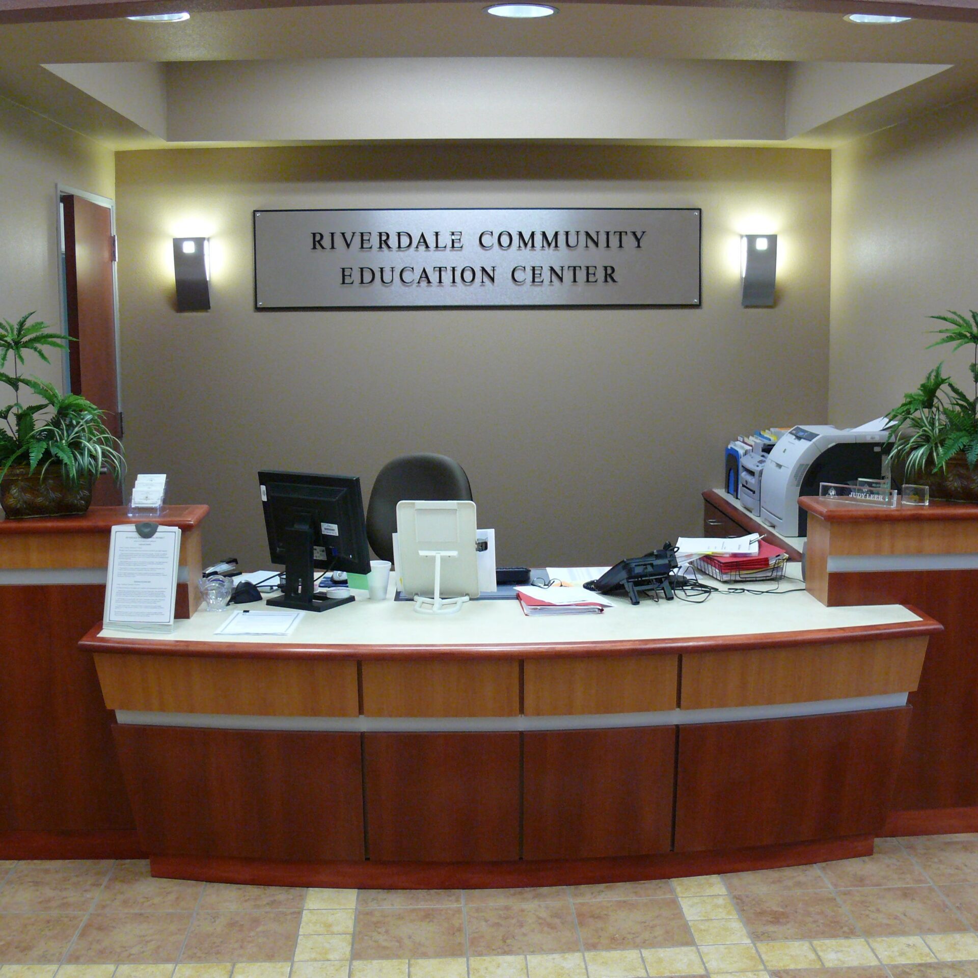 The interior of a building's reception area. A curved, wooden front desk is in the center, with a computer monitor, phone, and other office equipment. Behind the desk, a sign reads "RIVERDALE COMMUNITY EDUCATION CENTER." Two potted plants sit on either side of the desk, and wall sconces provide light. The floor is tiled in a checkered pattern.