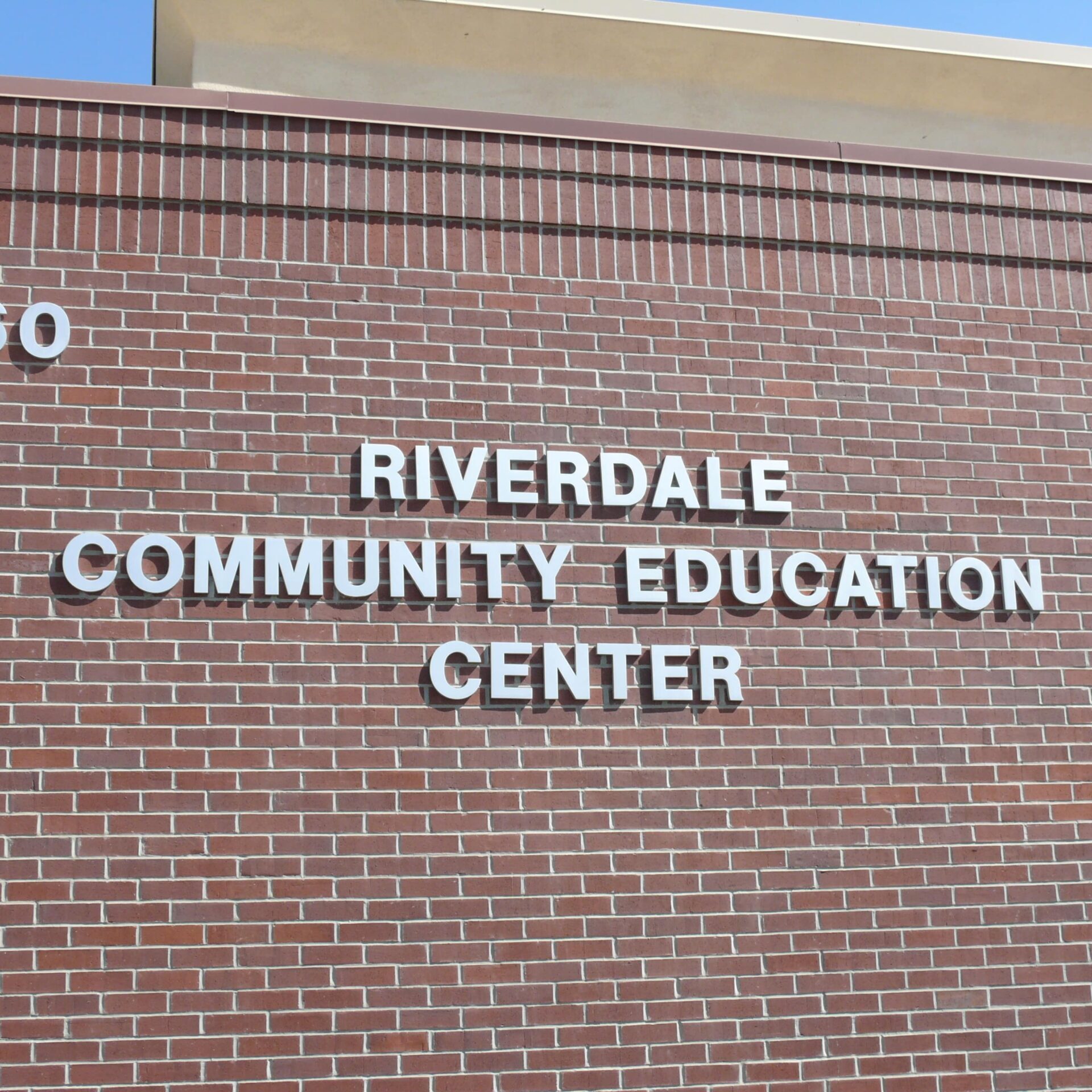 A close-up shot of a modern brick building. The building number '3160' is in the top left corner. The words "RIVERDALE COMMUNITY EDUCATION CENTER" are in the center, spelled out in large, white, raised letters.