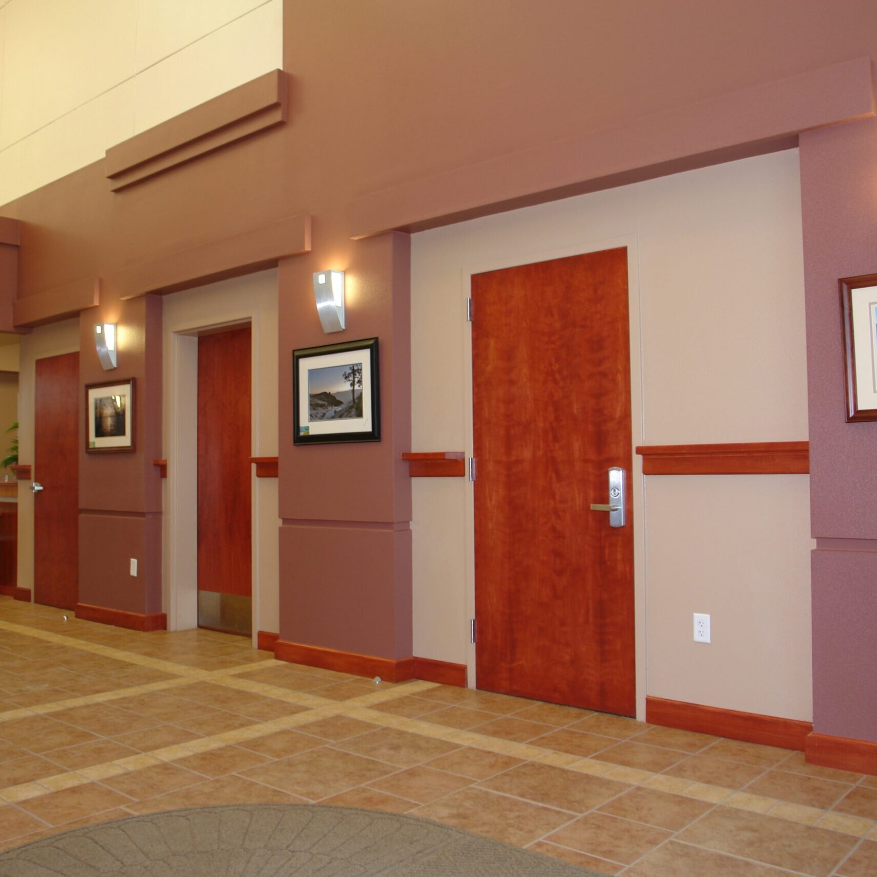 The interior hallway of a modern building. There are multiple wooden doors and framed art on the walls. The walls are painted in shades of brown and beige, and the floor is a checkered tile pattern. Light emanates from recessed ceiling lights and wall sconces.