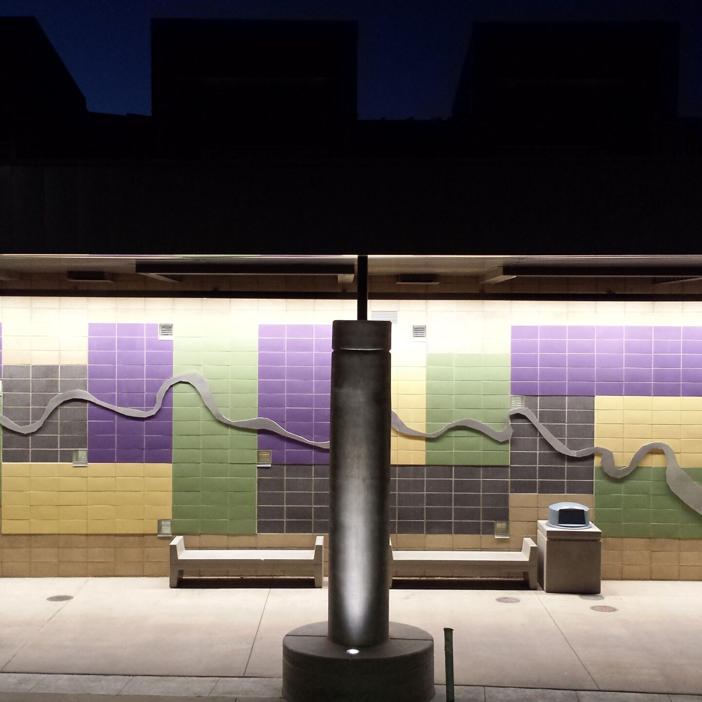 A public art installation on an exterior wall at night. The wall is covered in a grid of square tiles in varying shades of purple, light green, and yellow. A stainless steel relief sculpture of a winding river, representing the San Joaquin River, is mounted over the tiles. A bench and two round picnic tables are in the foreground. The scene is illuminated by an overhead light fixture.