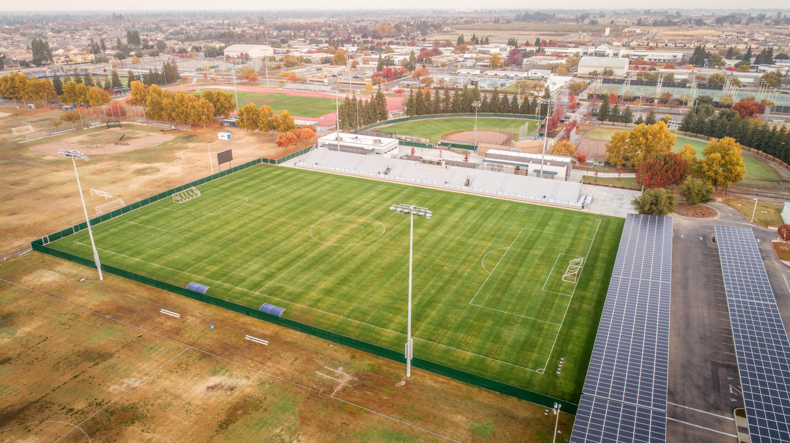 The Clovis East Soccer Stadium 000003-min An aerial view of the Clovis East High School campus, focusing on a sports complex. In the foreground, there is a large, well-maintained soccer field with bright green grass, surrounded by a low green wall. Tall stadium lights are positioned on the sidelines. To the right of the field, there are rows of solar panels. In the background, there is a baseball diamond, a track, and bleachers, surrounded by autumn-colored trees. The image captures a wider view of the surrounding residential neighborhood under a cloudy sky.