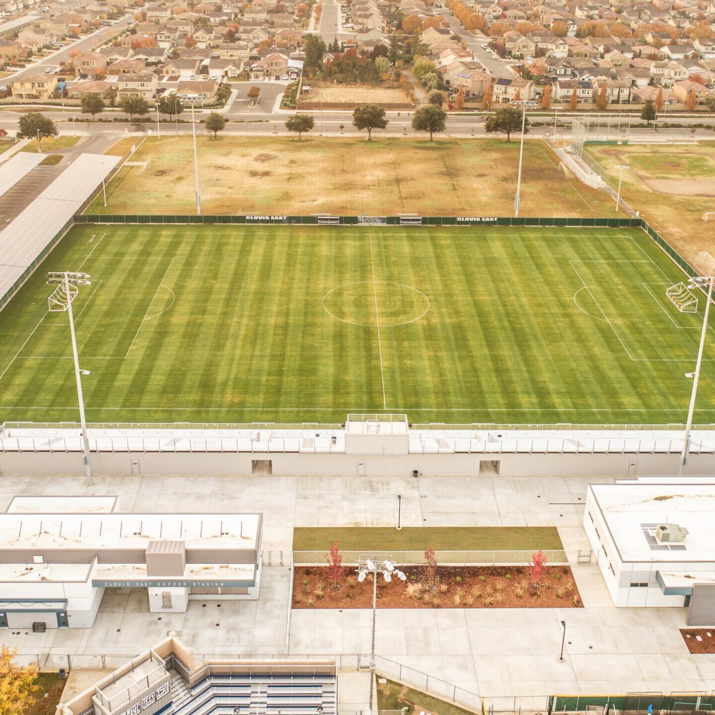 A wide aerial shot of the Clovis East Soccer Stadium and a residential neighborhood in the distance, showing the green field and bleachers.