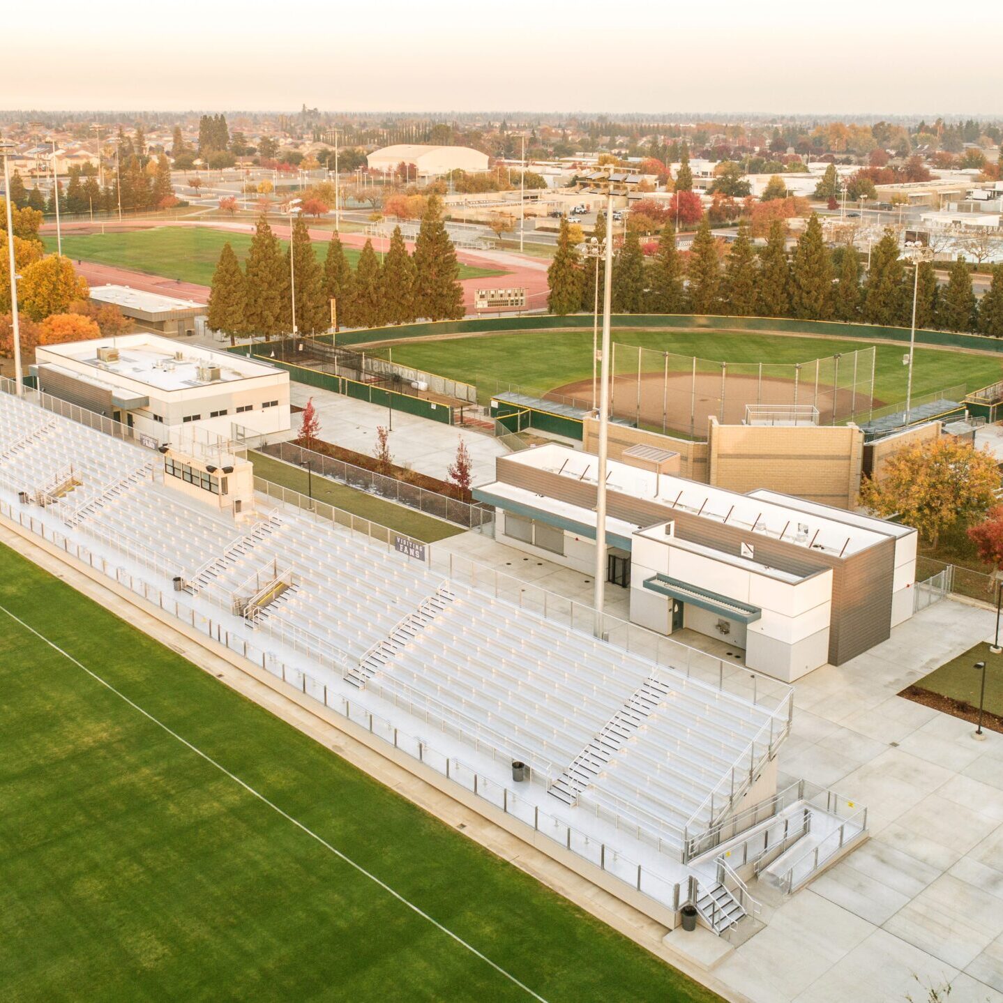A wide, high-angle view of the stadium's bleachers and two small buildings, with a baseball field and trees in the background.
