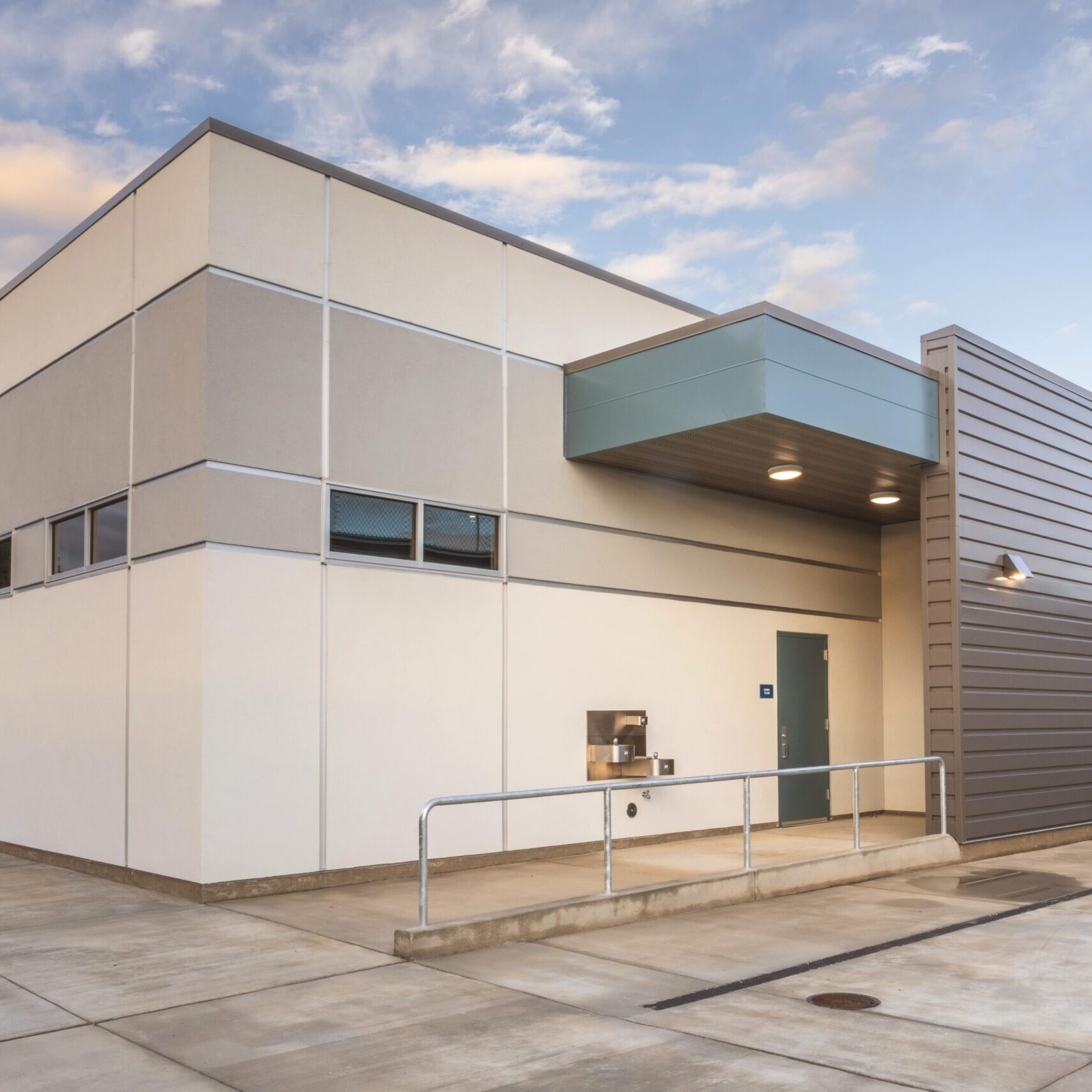 A ground-level shot of a building with a light-colored facade and a dark, corrugated metal section, showing a drinking fountain on the wall.