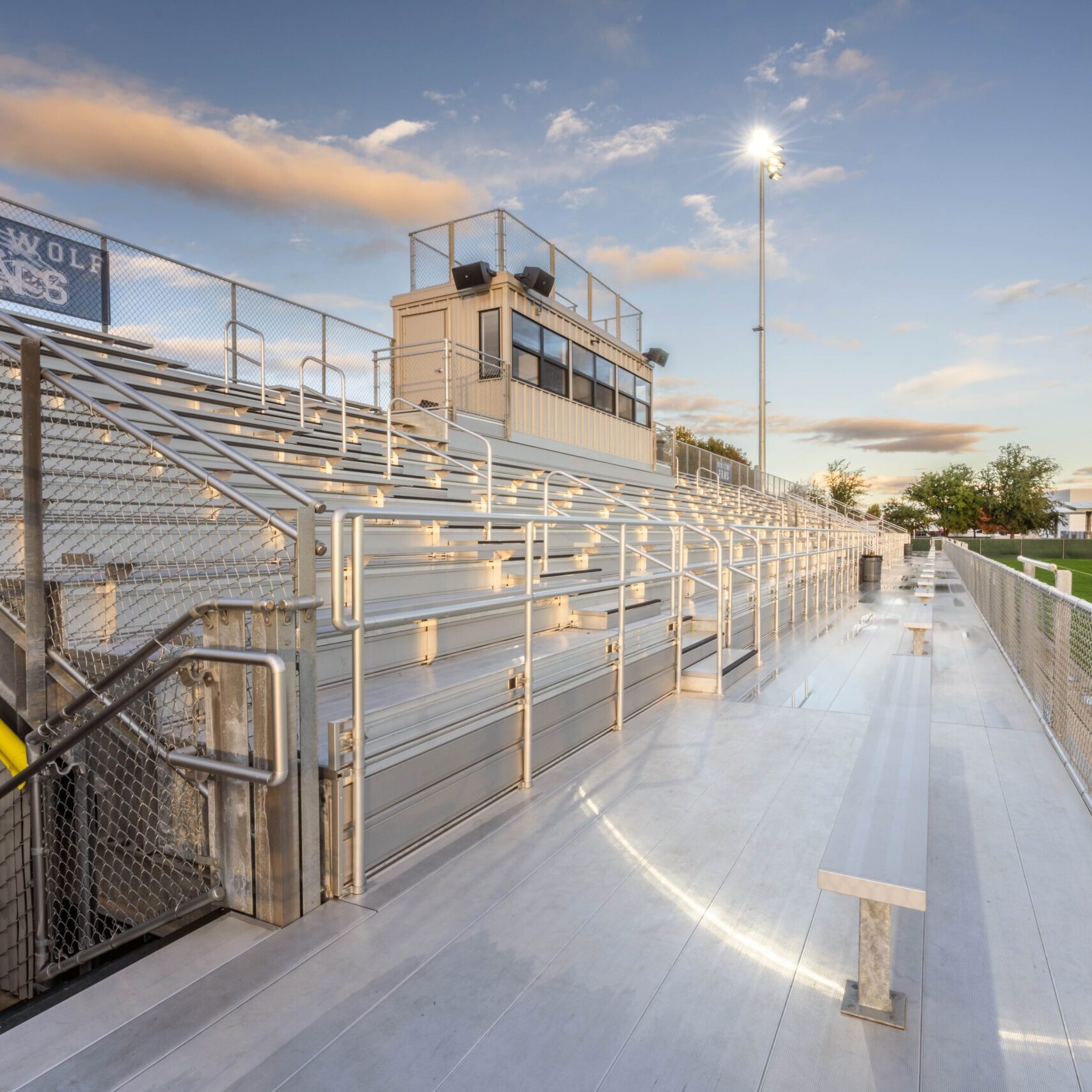 A low-angle shot of the stadium's metal bleachers and press box, with a sign that reads "TIMBERWOLF" and a bright light pole.