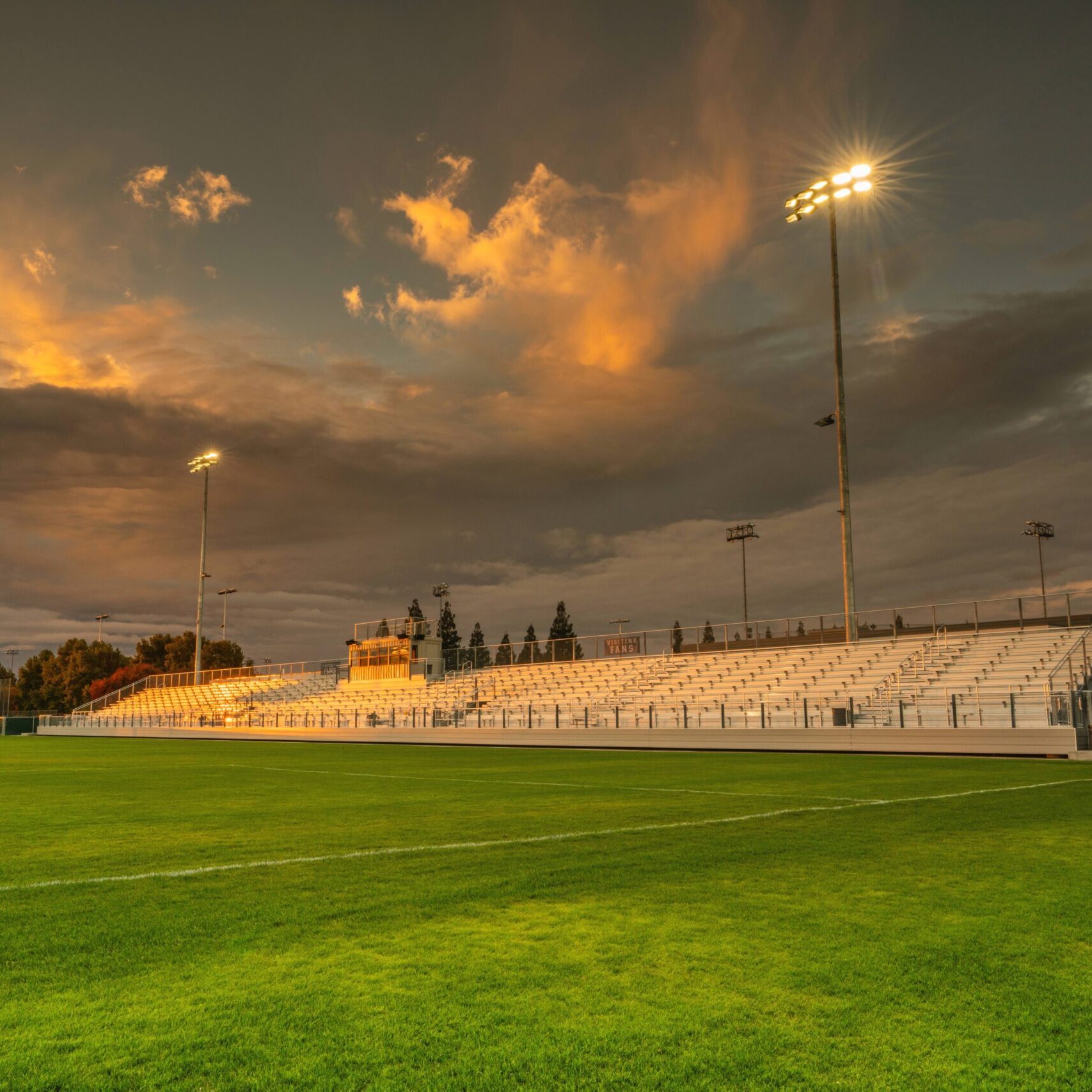 A wide, low-angle shot of the soccer field and bleachers, with a dramatic, cloud-filled sunset in the sky.