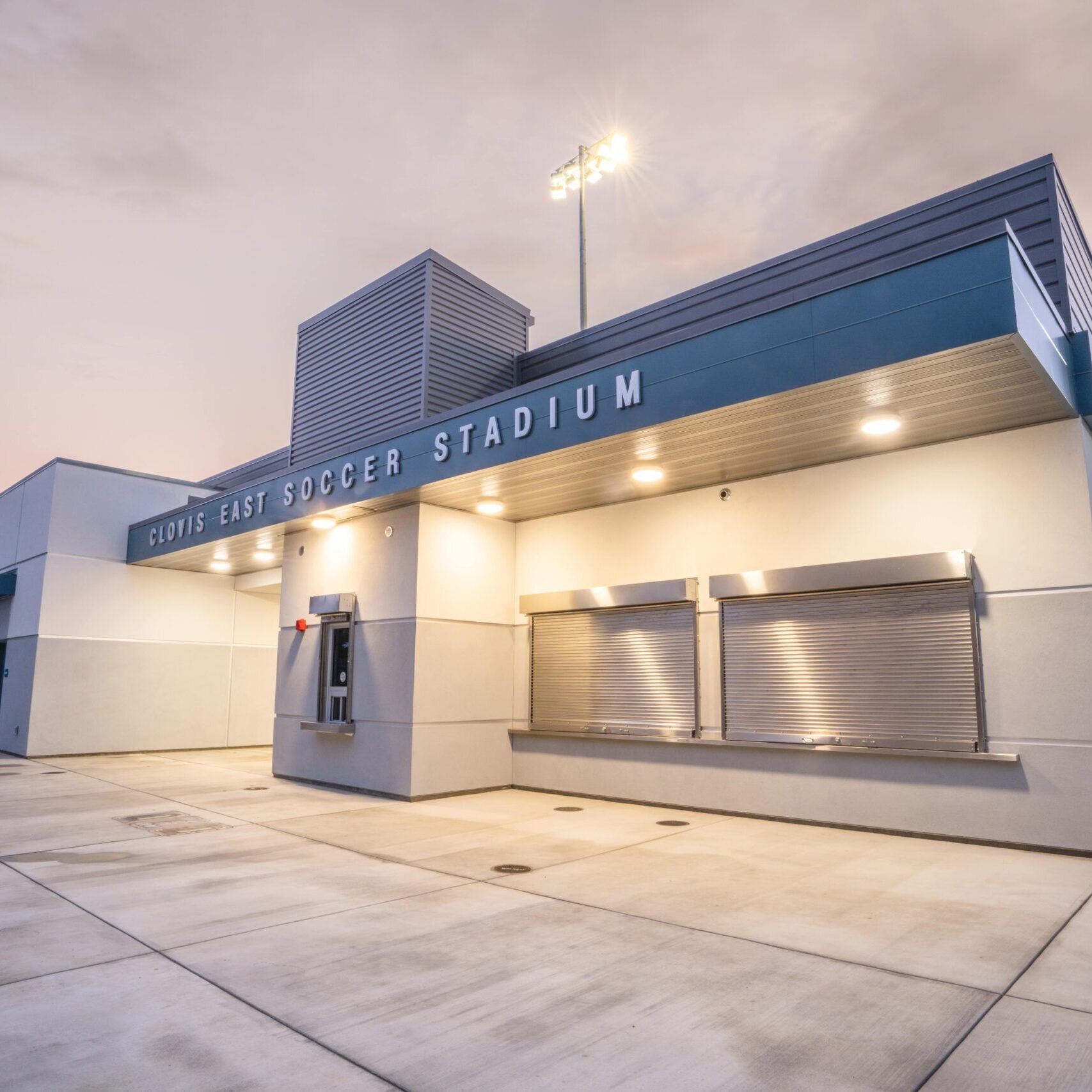A low-angle shot of the stadium's main building at dusk, with "CLOVIS EAST SOCCER STADIUM" visible on the facade and lights illuminating the building.