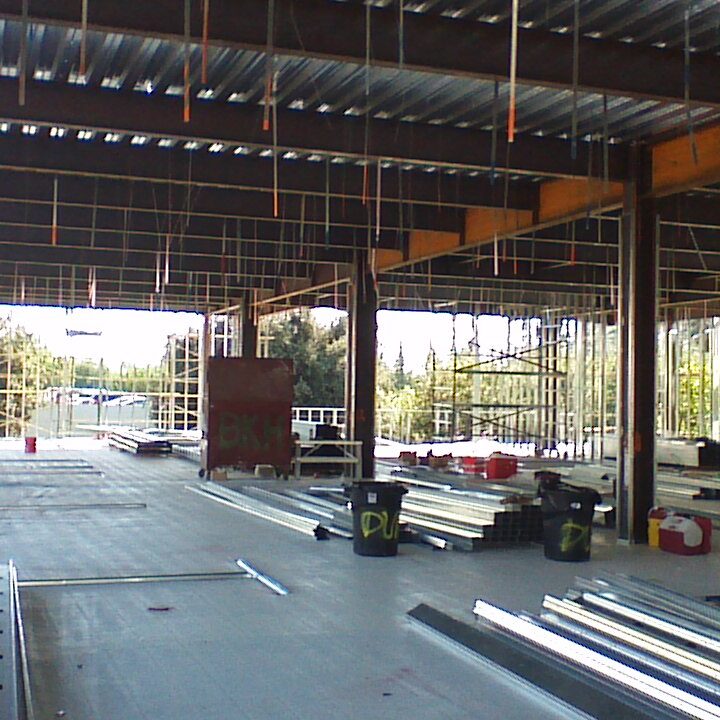 The interior of a large building under construction. The steel frame is exposed, with metal studs forming the interior walls. The ceiling has corrugated metal decking and exposed beams. Piles of steel studs and other building materials, along with black trash cans, are scattered across the concrete floor. Outside, a parking lot and trees are visible through the large window openings.