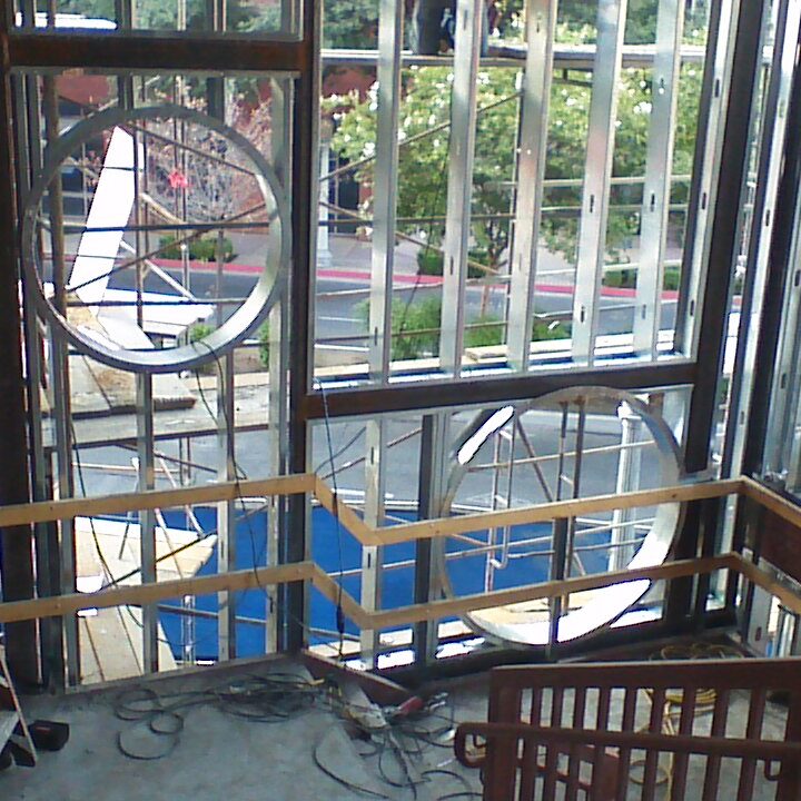 The interior of a building under construction shows a large, multi-story window area with exposed steel framing and metal studs. Two large, circular window frames are visible within the wall. A temporary wooden ladder is propped against a wall on the left, and a temporary wooden railing is visible along the edge of a balcony. A courtyard area with trees and paving is visible through the windows.