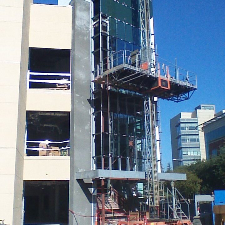 A multi-story building under construction is visible from a low angle. A vertical construction hoist is attached to the side of the building, which has a glass facade and dark green paneling. The adjacent part of the building is unfinished, showing square window openings and an unpainted concrete wall. In the foreground, there is a dirt path and construction equipment.