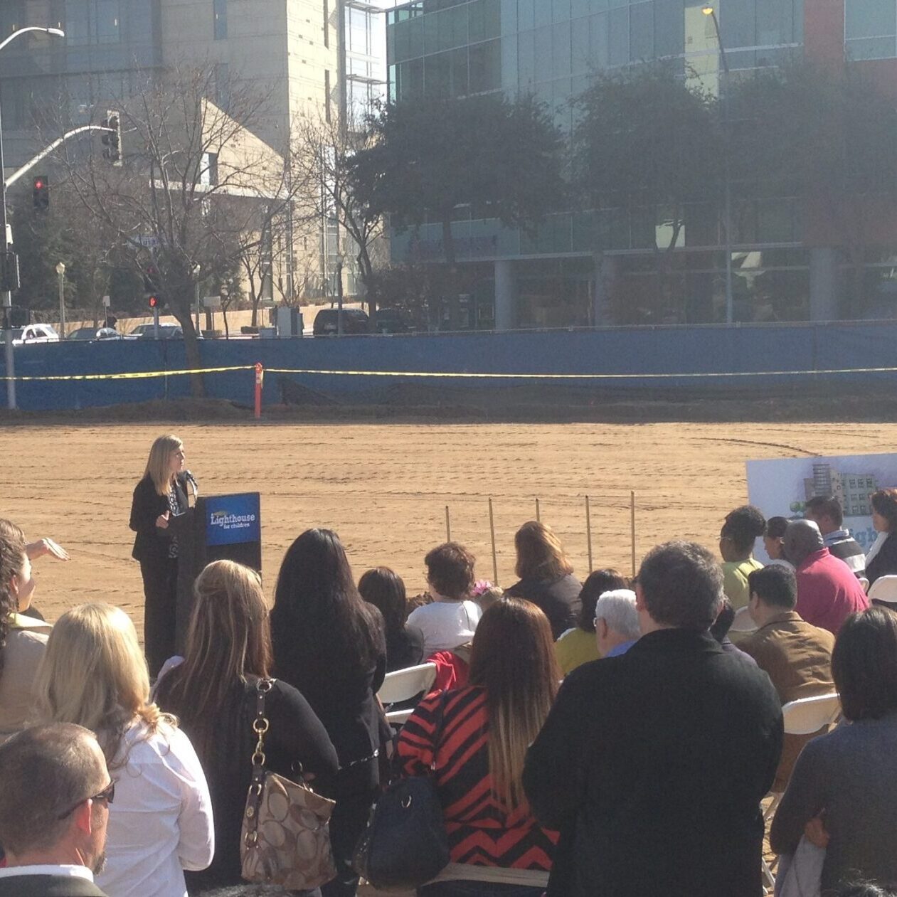 A woman is speaking at a podium on a large, empty dirt lot. A crowd of people is seated in the foreground, facing the speaker. A sign for the "Lighthouse for Children" is visible to the right of the podium. In the background, there are modern buildings and trees, with yellow caution tape visible along the fence line.