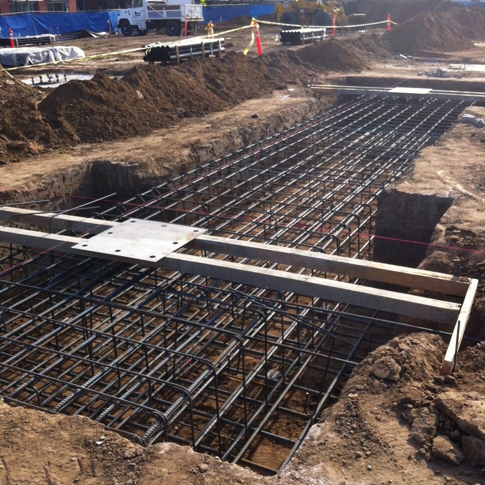 A deep foundation pit on a construction site shows a large, rectangular grid of steel rebar cages in place, ready for a concrete pour. The surrounding dirt is piled up, and wooden formwork and a metal plate are laid across the rebar. In the background, there is a large yellow excavator, a truck, and other urban buildings.