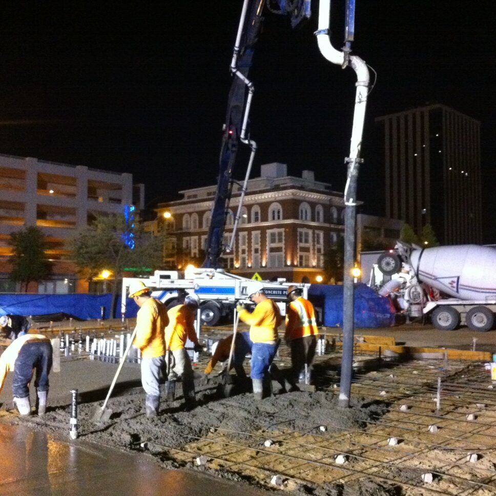 A nighttime construction scene shows a large concrete pouring operation. Several workers in hard hats and reflective vests are using shovels and tools to spread freshly poured concrete. A long, white concrete pump pipe extends from a truck, and a large concrete mixer truck is also visible in the background. The site is illuminated by bright lights, with city buildings visible behind a temporary fence.