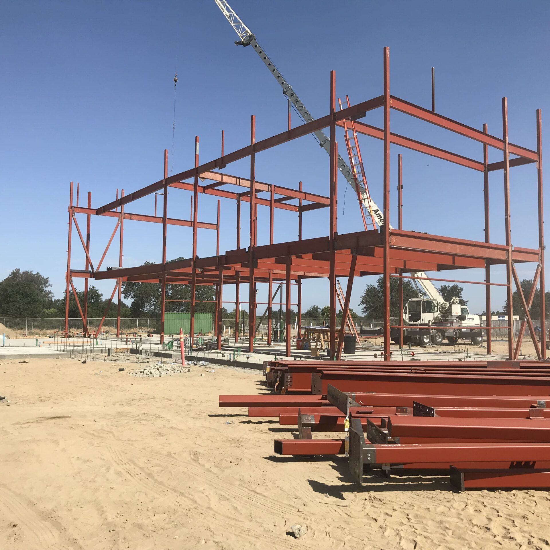 A ground-level shot of a construction site with the red steel frame of a two-story building partially erected. A large pile of red steel beams is in the foreground, and a construction crane is visible in the background.